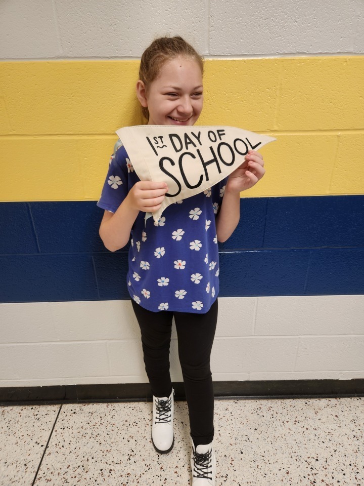 A female student stands in front of a blue and yellow stripped wall holding a sign that says "First Day of School."