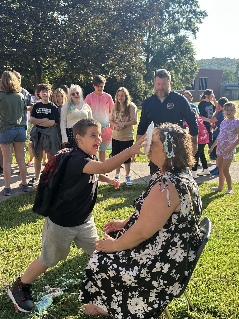 A male student excitedly throws a plate of pie in Mrs. Hesse's face. A crowd of students observe in the background.