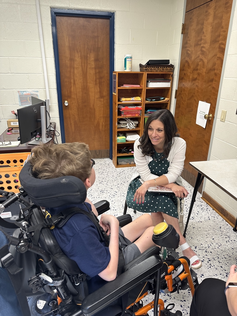 Ms. Ashley Suddath is seated with a book on her lap as she smiles at a male student in a wheelchair.