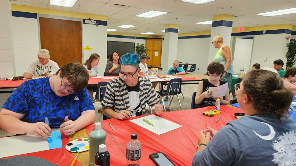 Students seated at a long, rectangular table full of art supplies as they work on their art activity.