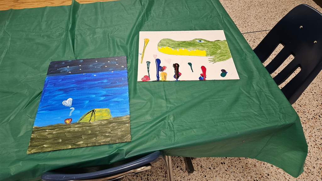 Two finished art activities sitting on a table for display. The art on the left is a camping trip with a tent and starry night, while the art on the right is a green alligator.