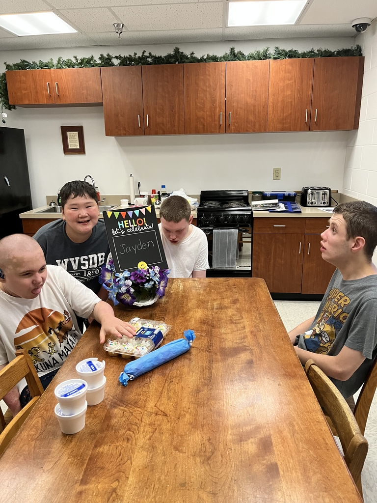 Students seated at a wooden table with cupcakes and ice cream. One student stands and holds up a birthday sign for one of their fellow students who is seated.