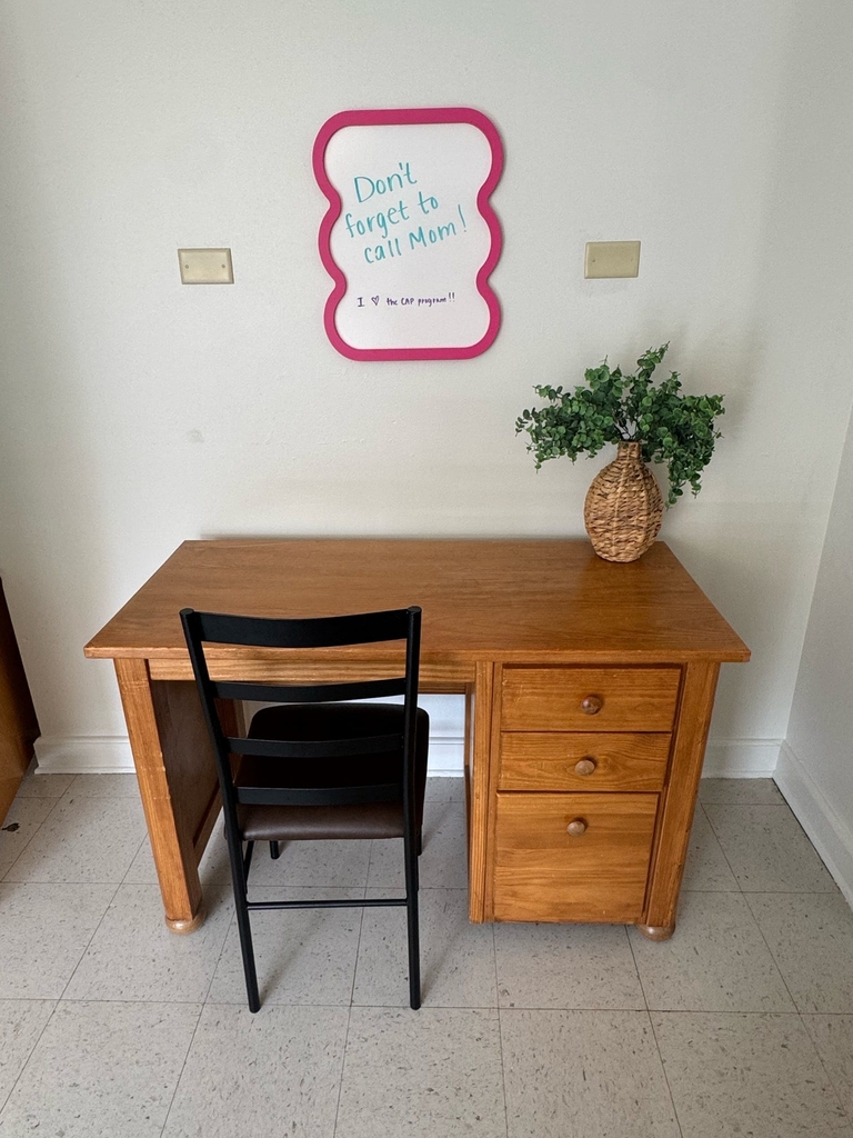 Staged CAP dorm room featuring a wooden desk and chair with a green plant on top of the desk. A sign above the desk reads "Don't forget to call mom."