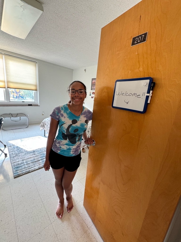 A female CAP student holds open her dorm room door while she poses for the camera.