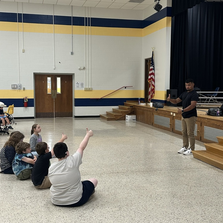 Students seated on the floor give a “thumbs up” to Justin Perez, who is standing in front of the audience.