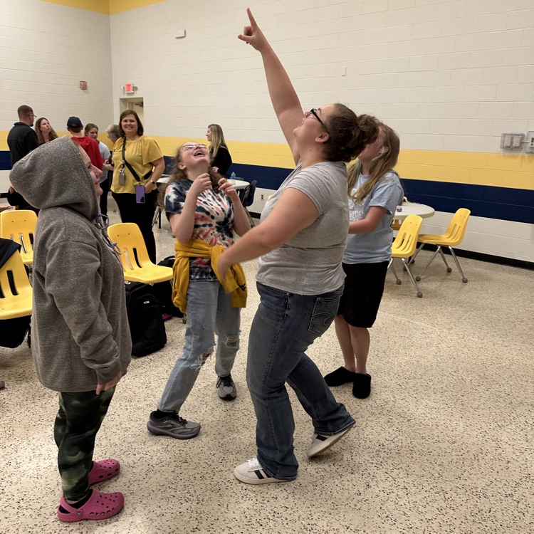 A group of students act out a scene during an activity. A female student points to the ceiling and smiles.