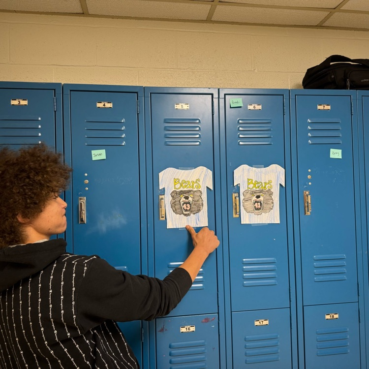 Daevon, a CAP student points to a paper jersey with a black bear on it as it hangs on a blue-colored locker.