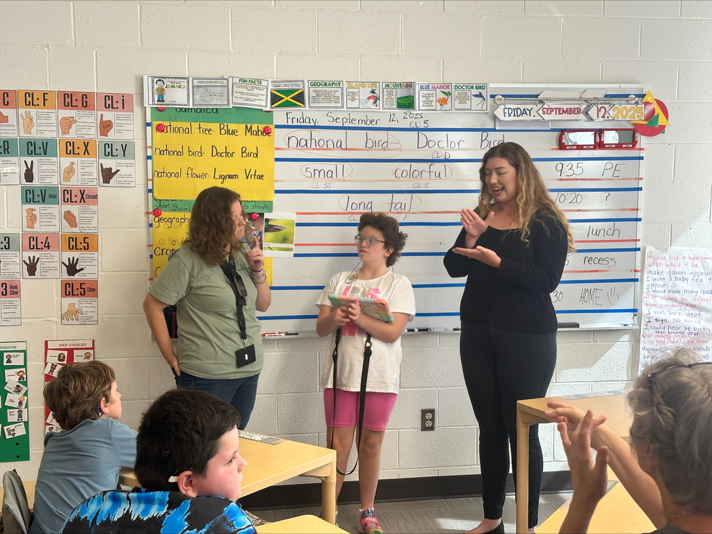 Mrs. Shoemaker stands at the front of the classroom with a female student and a female ASL interpreter.