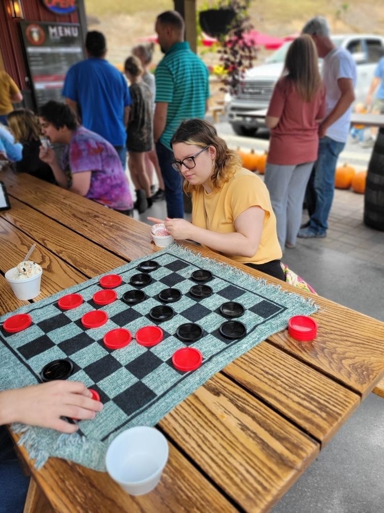 A female student looks down as she scoops her ice cream to eat. There is a game of checkers sitting in front of her.