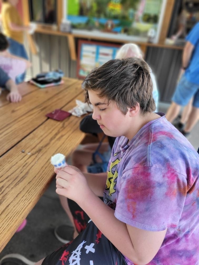 A male student seated at a wooden picnic table looks down at his ice cream cone.
