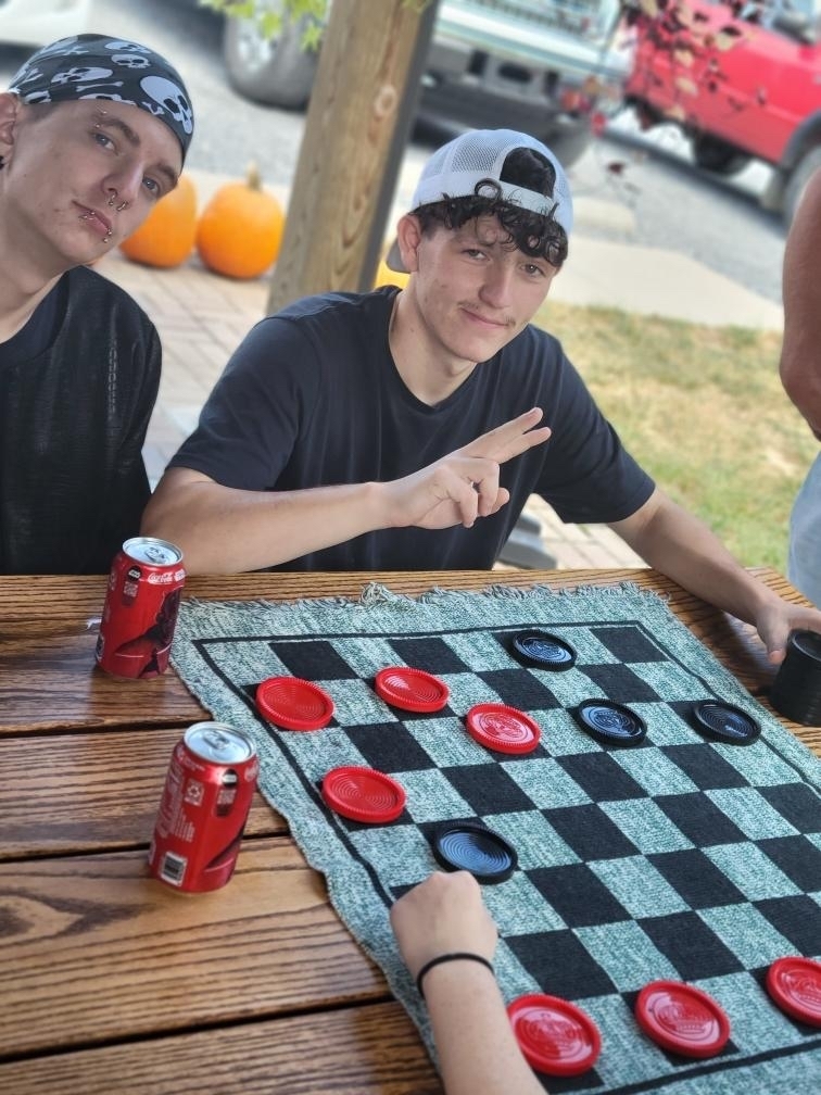 Two male students seated side by side on a wooden bench with a game of checkers in front of them.