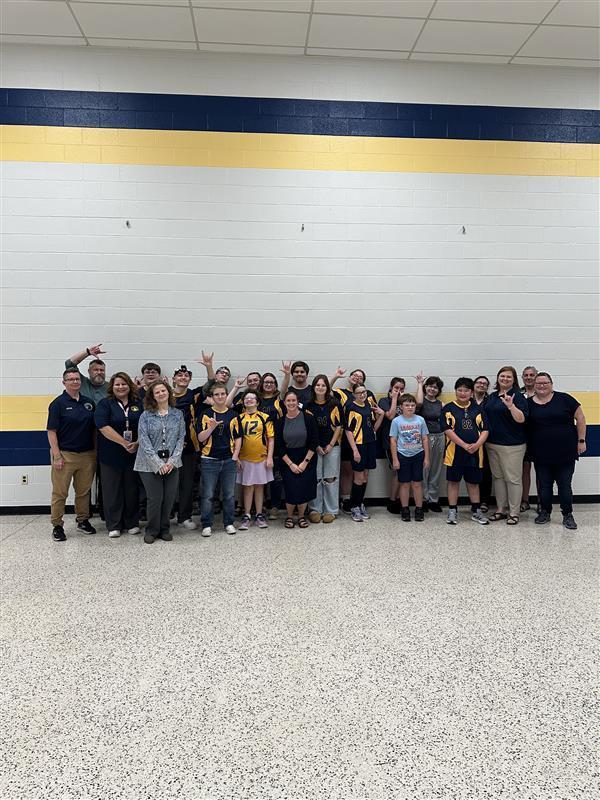Students and staff stand side by side in a group, some holding up the "I love you" sign in ASL in front of a wall with blue and yellow stripes.
