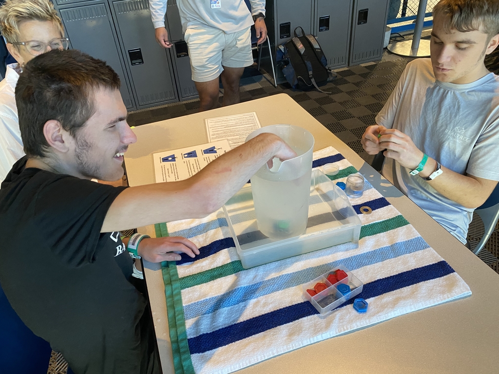A male student is seated with another male student as they engage in a STEM activity using water.