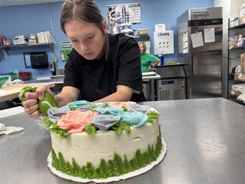 A female student looks down as she puts green-colored icing onto a white cake.