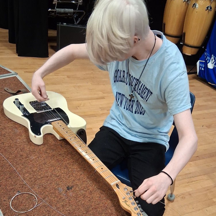 A male student looks down as he strings a beige-colored electric guitar.