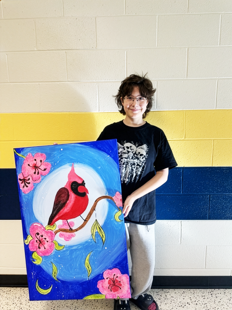 A female student holds up a large canvas painting of a red cardinal with pink flowers surrounding the cardinal.