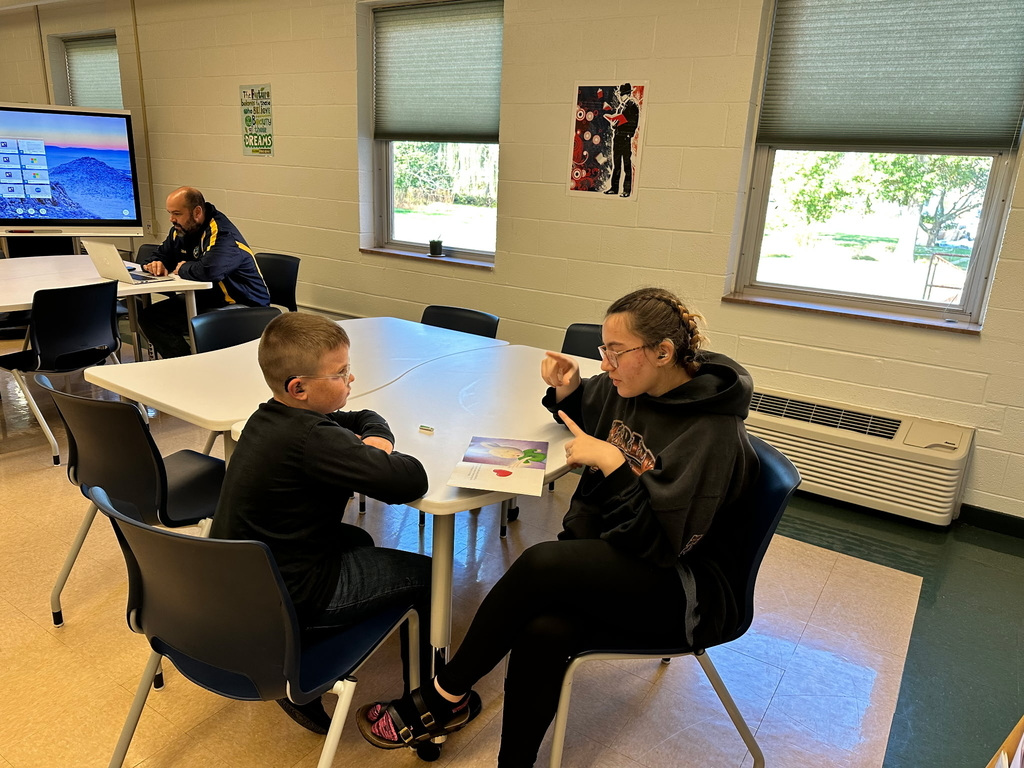Two students seated at a table. The female student assists a male student with his math work.