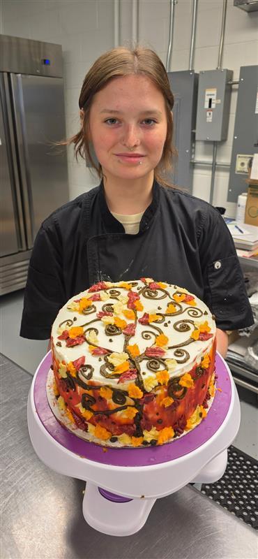A female student in a black culinary jacket holds a round-shaped cake that is decorated with orange, red, and brown accents.