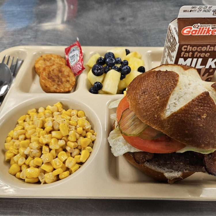 Meal tray featuring a hamburger with toppings, fresh fruit, corn, and chocolate milk.