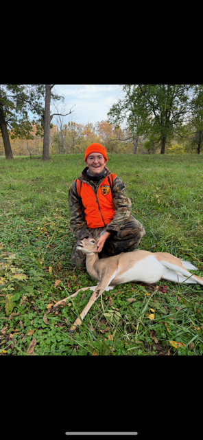 A male student wearing hunting gear proudly poses with a deer.