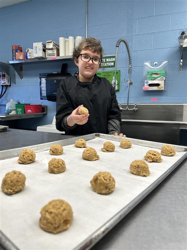 A female student dressed in a black culinary uniform smiles for the camera as she holds up cookie dough in her hand.