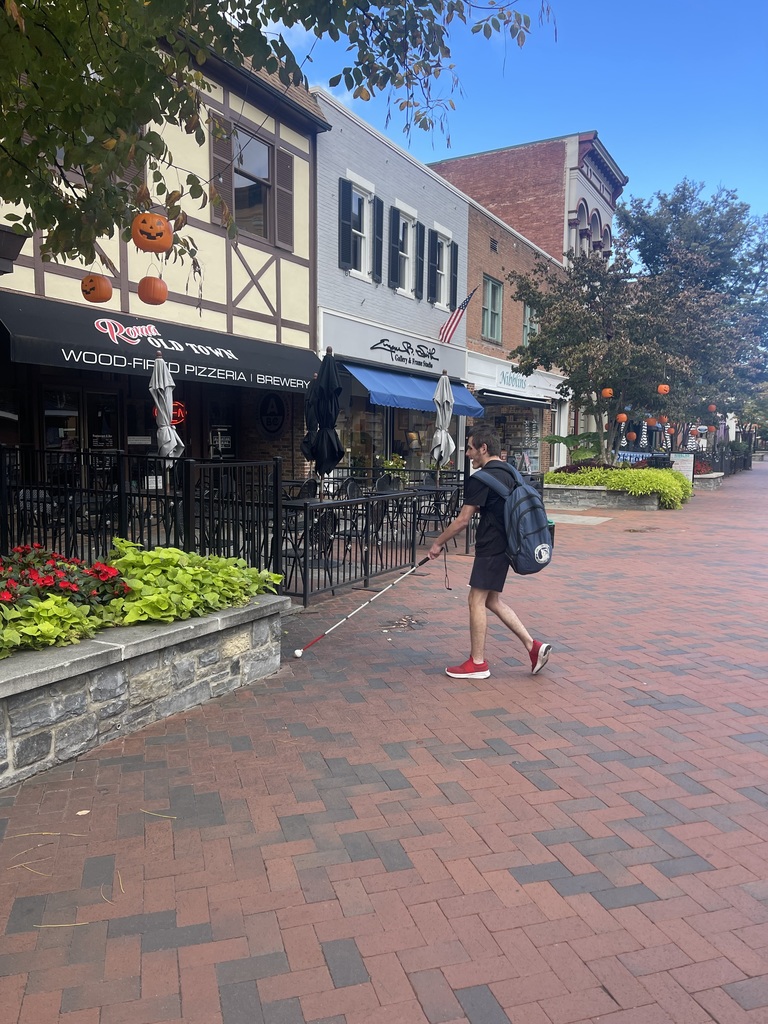 A male student uses his white cane to help guide him to the entrance of an Italian restaurant.