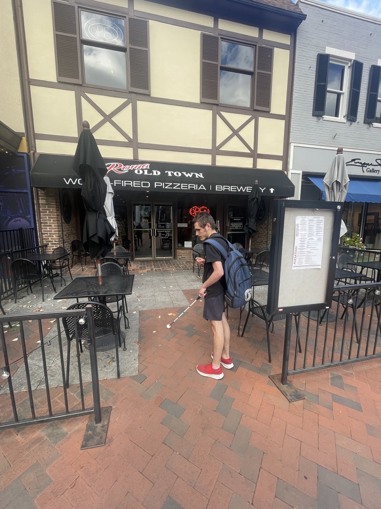 A male student uses his white cane to help guide him to the entrance of an Italian restaurant.
