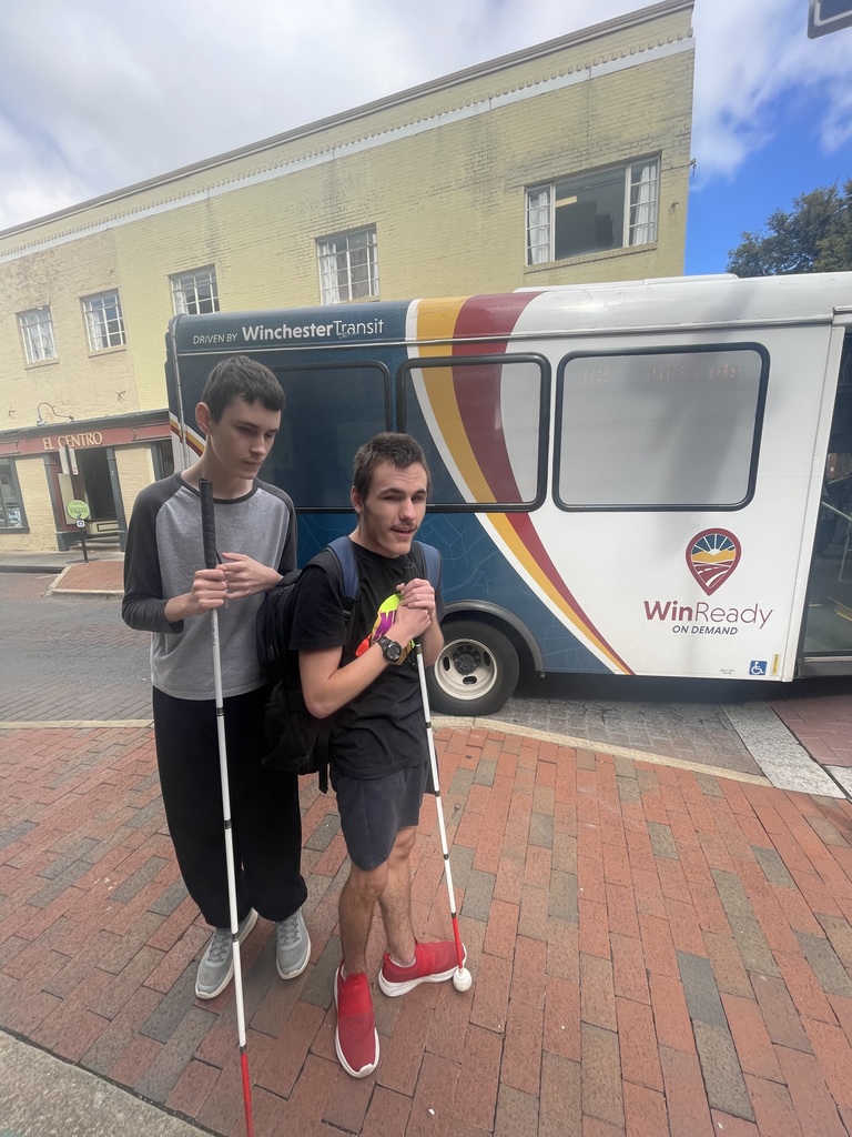 Two male students stand side-by-side with their white canes, standing on a sidewalk. There is a bus in the background behind them.