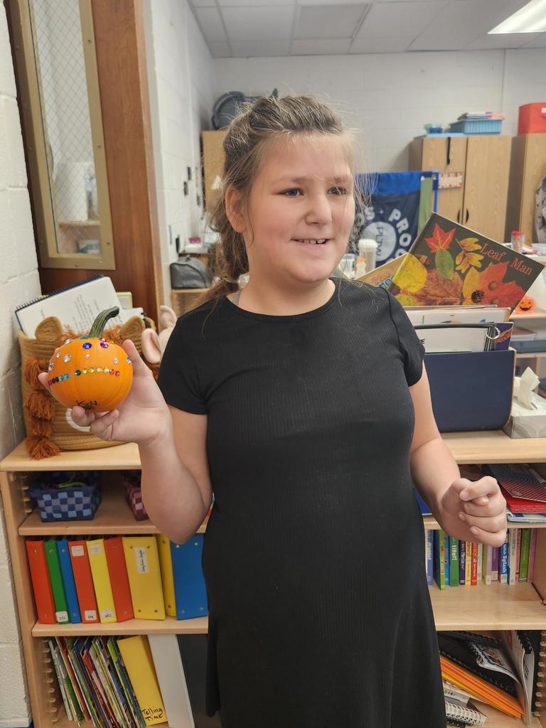 A female student with brown hair smiles as she holds up her orange, decorated pumpkin.