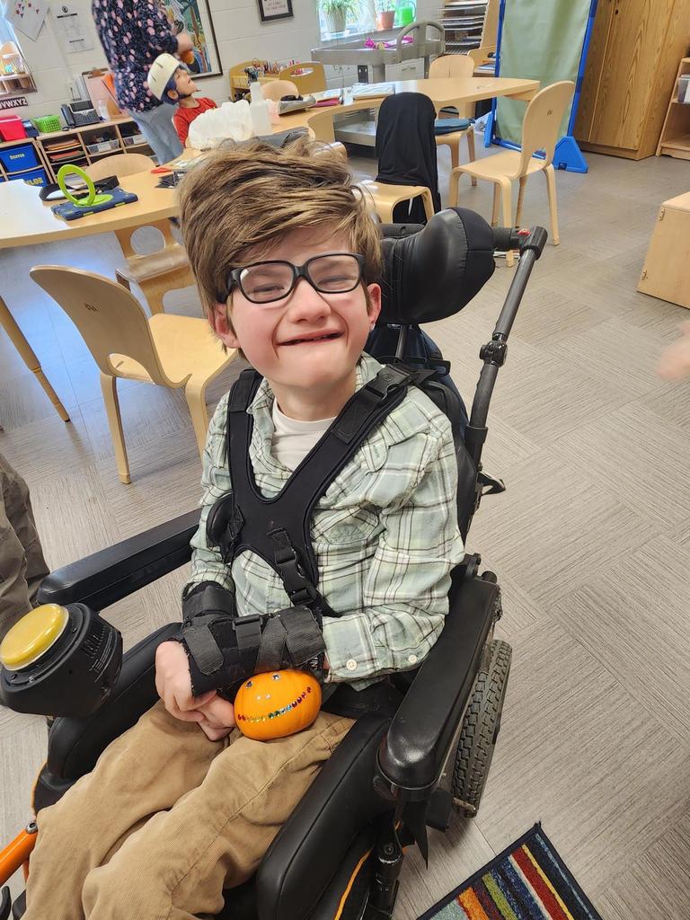 A male student in a wheelchair smiles at the camera as he holds his orange, decorated pumpkin.