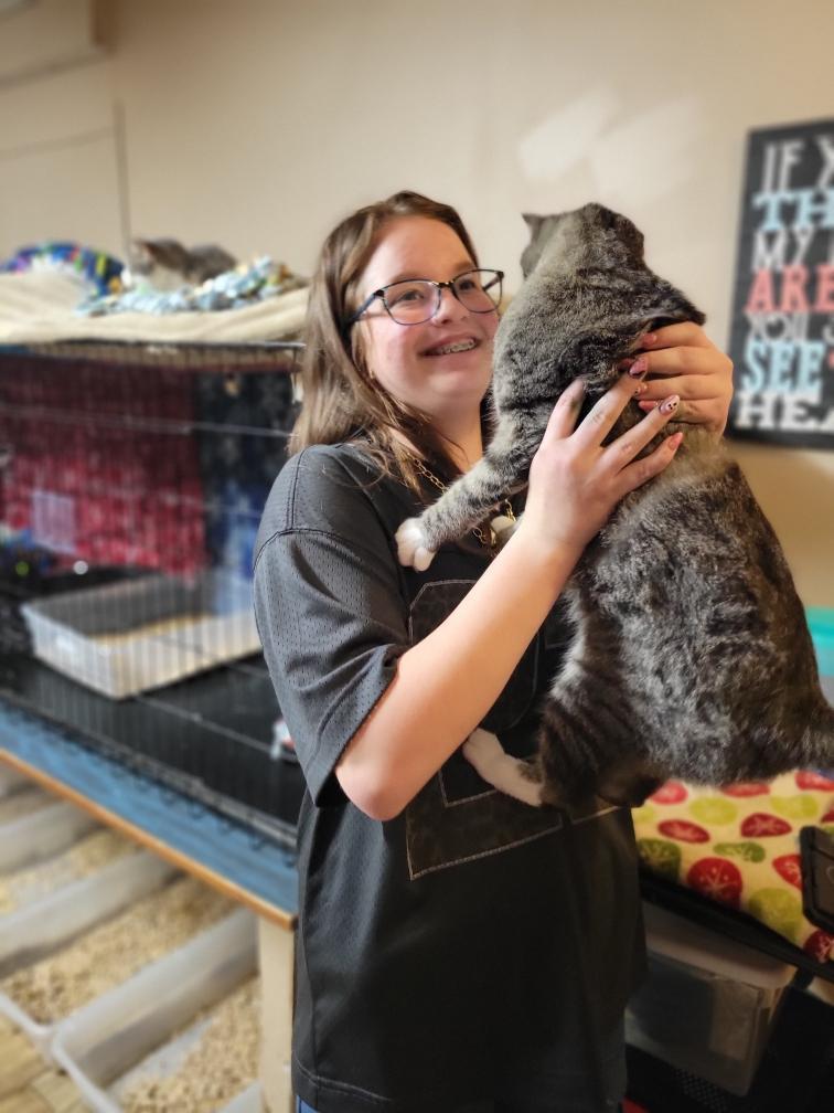 A female with brown hair and wearing glasses smiles as she holds up a cat in her hands.