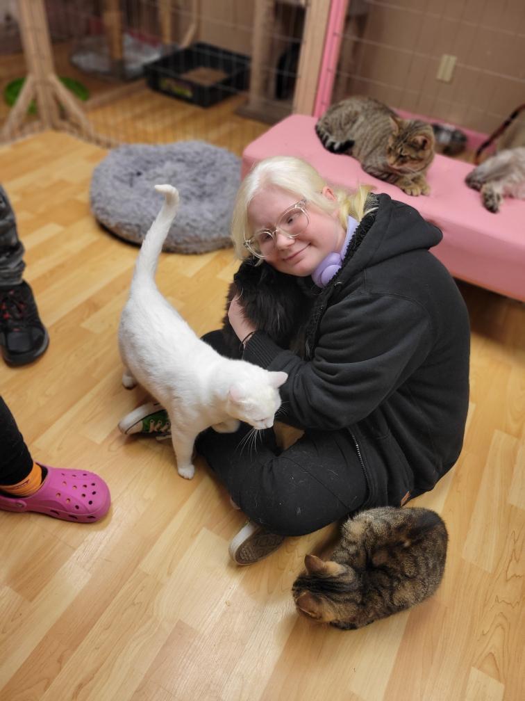 A female student with blonde hair and glasses is seated on the floor as she is surrounded by cats.