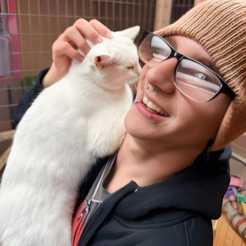 A male student wearing glasses and a beanie smiles as he holds a white cat in his arms.
