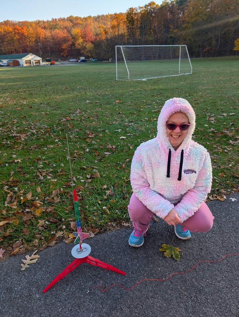 A female student wearing glasses and a pink fuzzy jacket with the hood up leans down to pose with her rocket that is on the launchpad.
