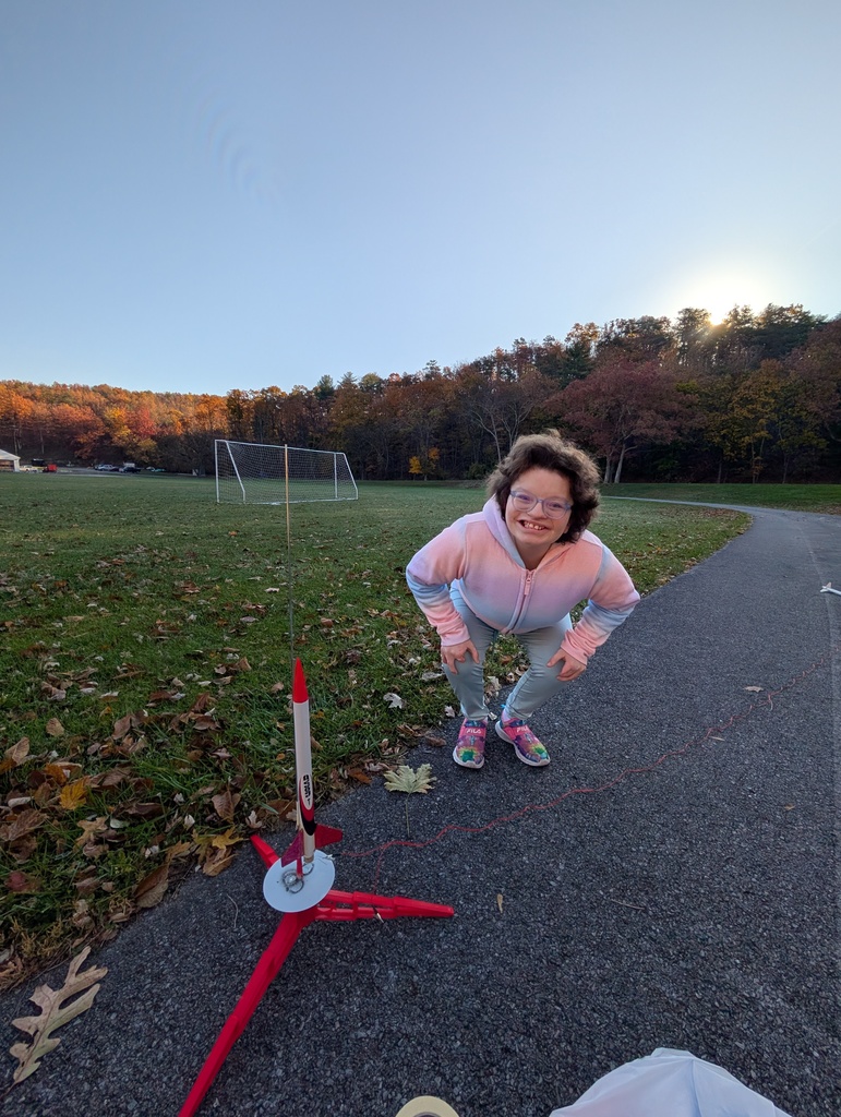 A female student with glasses and dark, curly hair smiles and poses with her rocket that is on the launchpad.