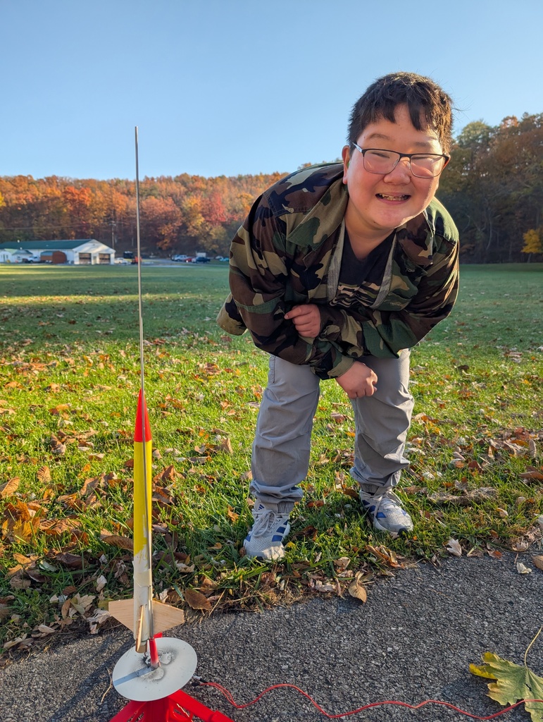 A male student with dark hair and glasses leans down to pose with the rocket he built.