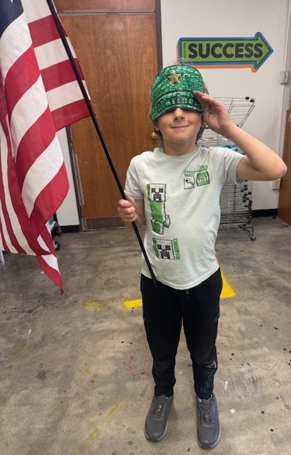 A male student stands proudly and salutes with one hand and holds up a red, white, and blue American flag with the other.