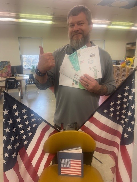 Roy, a male interpreter does a thumbs up and holds up cards that were created for Veterans Day. Two red, white, and blue American flags are positioned in front of him. 