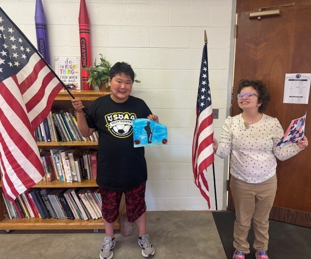 Two students hold up their Veterans Day-themed crafts they created in one hand, and they hold up a red, white, and blue American flag in the other.