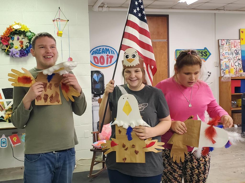 Three students hold up crafts they made to honor Veterans Day. A female student in the middle holds up a red, white, and blue American flag.