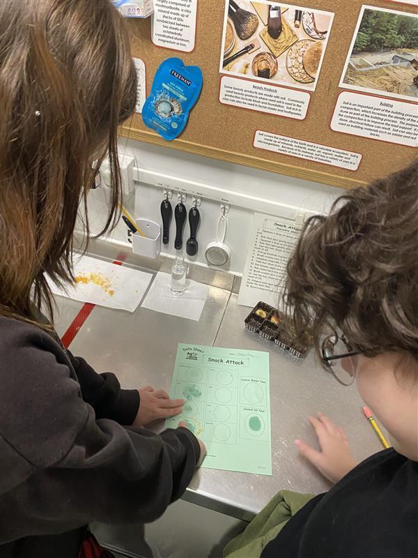 Two students look down as they fill out their green-colored science sheet during their experiment.