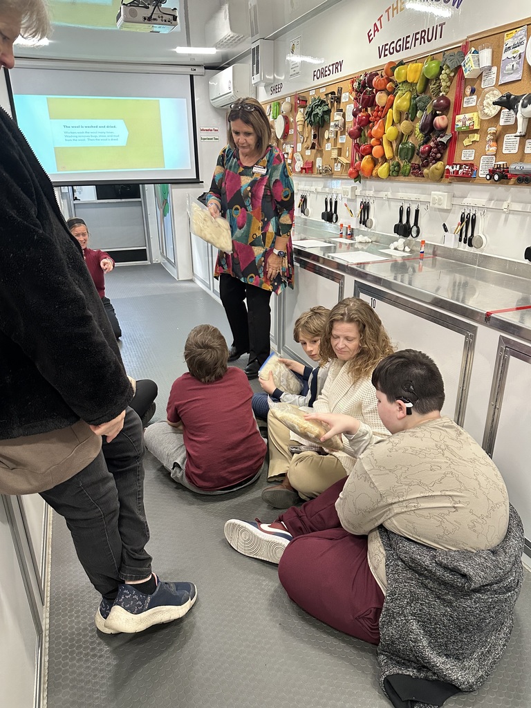 Students are seated on the floor as the female representative presents a presentation on the science lesson they are learning.