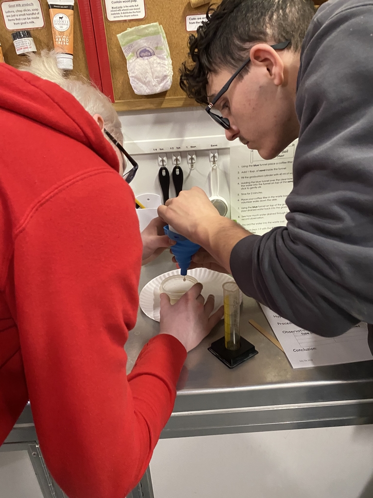 Two male students look down and concentrate as they put a substance into a blue-colored funnel.