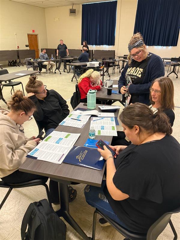 A group of attendees are seated at a rectangular-shaped table with resources sitting on the table in front of them.