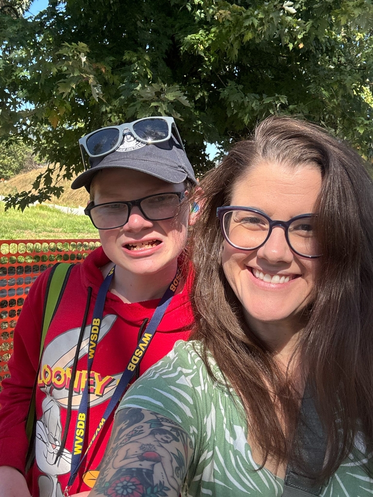 Ms. Rachel Lanham, a woman with dark hair and wears glasses takes a selfie with a male student who is wearing a hat and glasses.