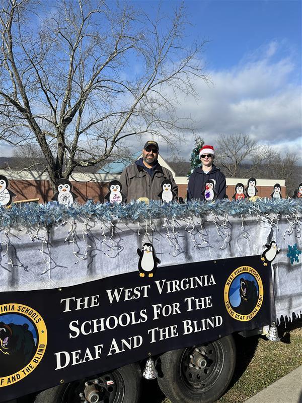 Mr. Haza, a male music teacher and a male student side by side while on the float. The float is decorated with blue and white tinsel, a WVSDB banner, and penguins.