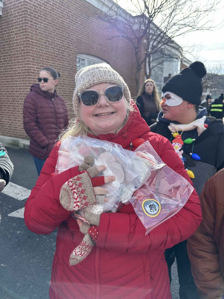 A female student wearing a bright, red jacket smiles as she holds packs of hot chocolate in her arms.