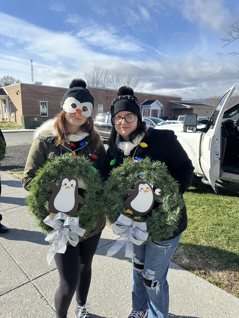 Two female students pose for the camera holding their green wreaths with a black and white penguin in the center of the wreath.