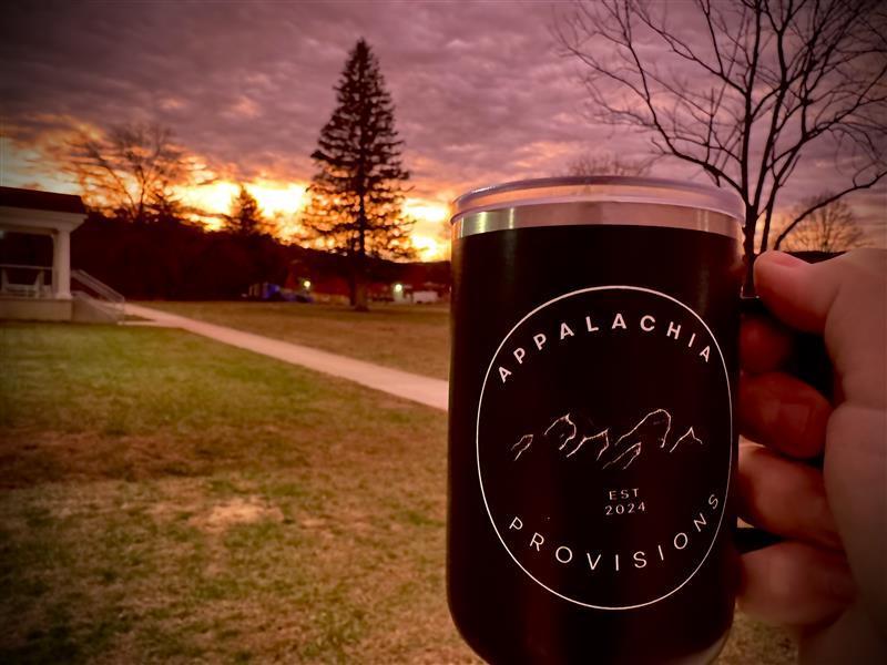 A black mug with the Appalachia Provisions logo on it is being held up in front of an orange sunrise.