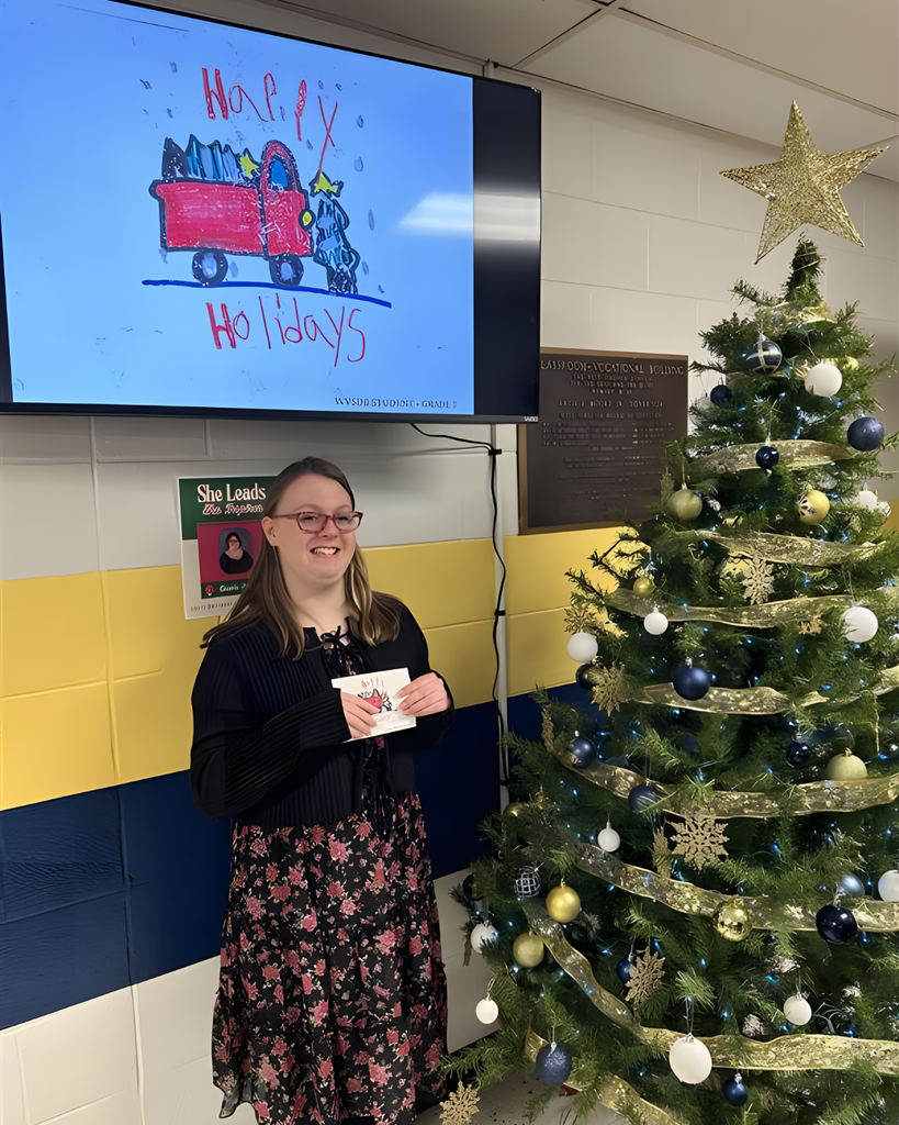 A female student wearing a black top and floral skirt smiles proudly as she holds up a Holiday card she created. On the TV display behind her, is her creation. A red-colored truck hauling a green tree. "Happy Holidays" is in red-colored font. There is a tree decorated with blue, white, and gold decorations to the right of the female student.